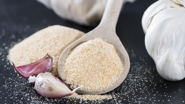 Garlic powder in a wooden spoon, next to garlic cloves