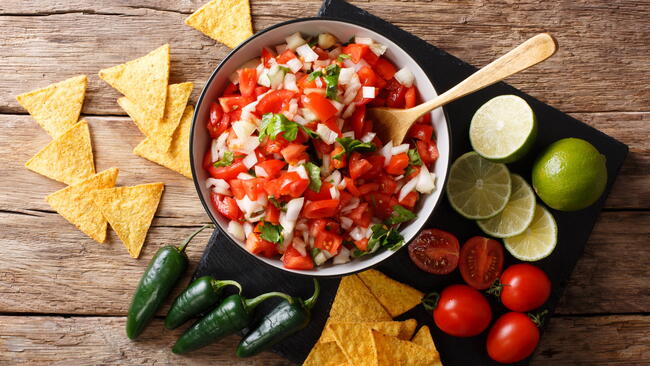A bowl of salsa surrounded by chips, cherry tomatoes, jalapenos, and slices of lime 
