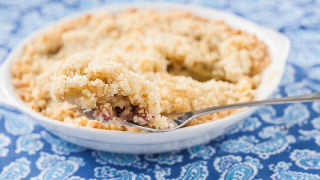 crumble with rhubarb and apple in a bowl on a blue tablecloth with a spoon
