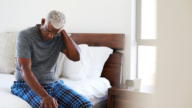 Senior Man Suffering With Neck Pain Sitting On Side Of Bed At Home