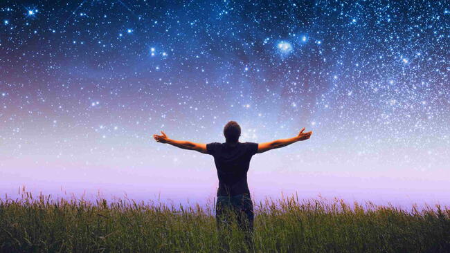 Person standing in a grassy field with arms outstretched under a starry night sky.