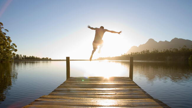 Horizontal rear view of a man leaping from a jetty into the lake at sunset.