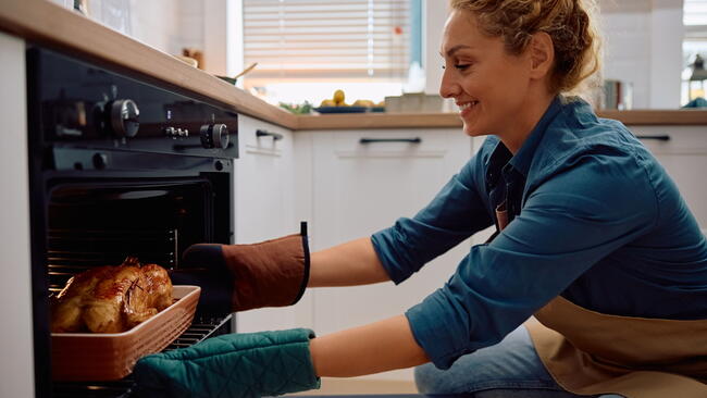 Happy woman taking roasted turkey out of the oven while preparing Thanksgiving dinner at home.