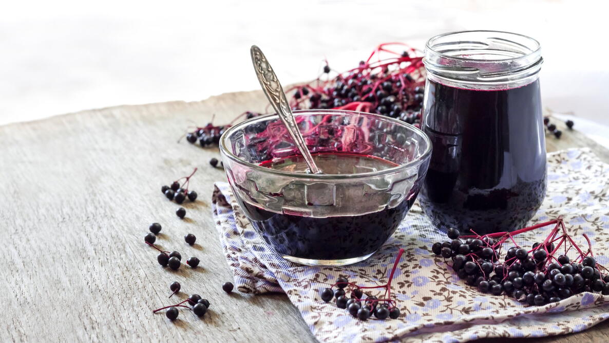  Homemade black elderberry syrup in glass bowl and jar and bunches of black elderberry in background.