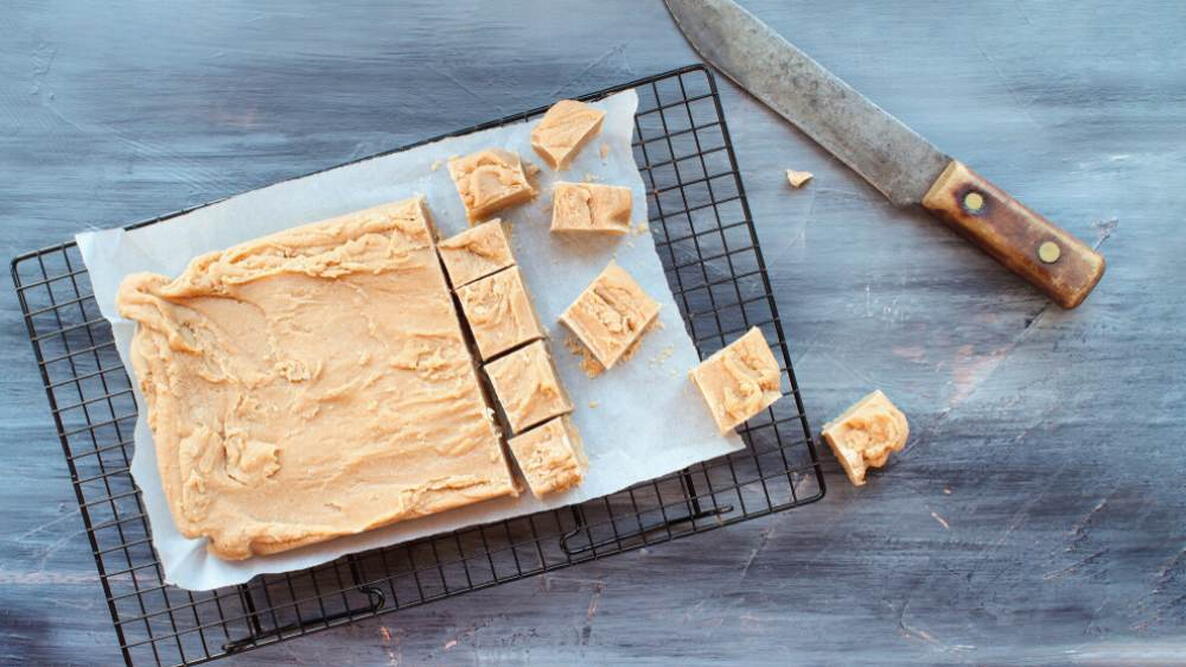 Block of delicious, homemade peanut butter fudge being cut into squares over a textured wood table background with old knife. Image shot from above.