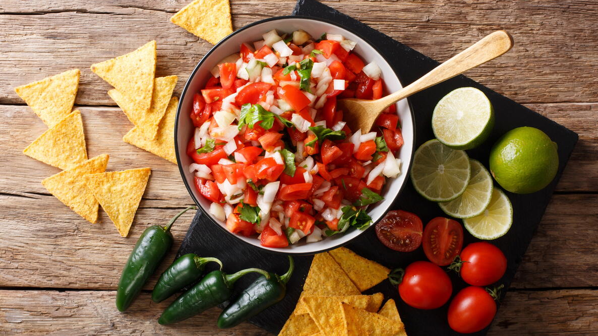 A bowl of salsa surrounded by chips, cherry tomatoes, jalapenos, and slices of lime 