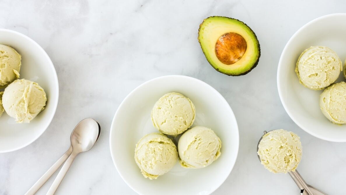 Avocado ice cream in three bowls, spoons, ice cream scoop and half an avocado on white marble 