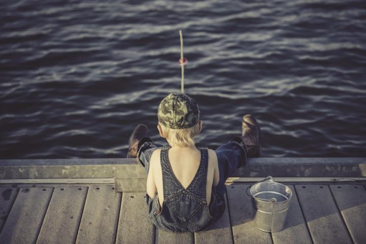 boy wearing overalls fishing off of the dock with a metal bucket beside him